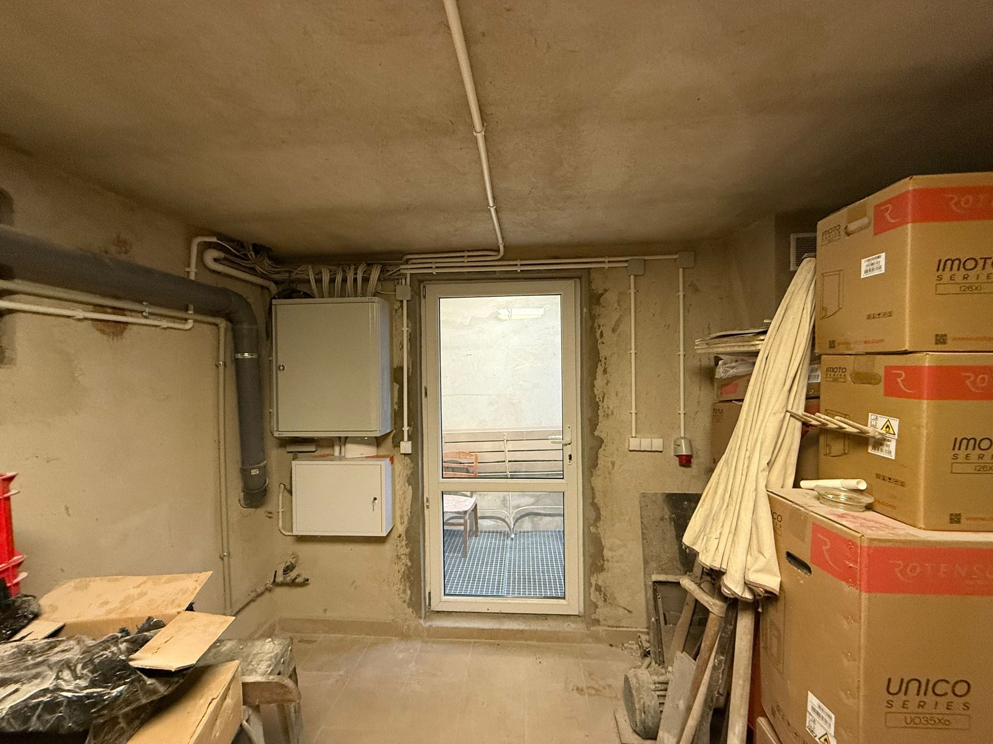 Piwnica z instalacją elektryczną, skrzynkami rozdzielczymi na ścianie, rurami na suficie i oknem na zewnątrz, obok okna składowane kartony i inne przedmioty.