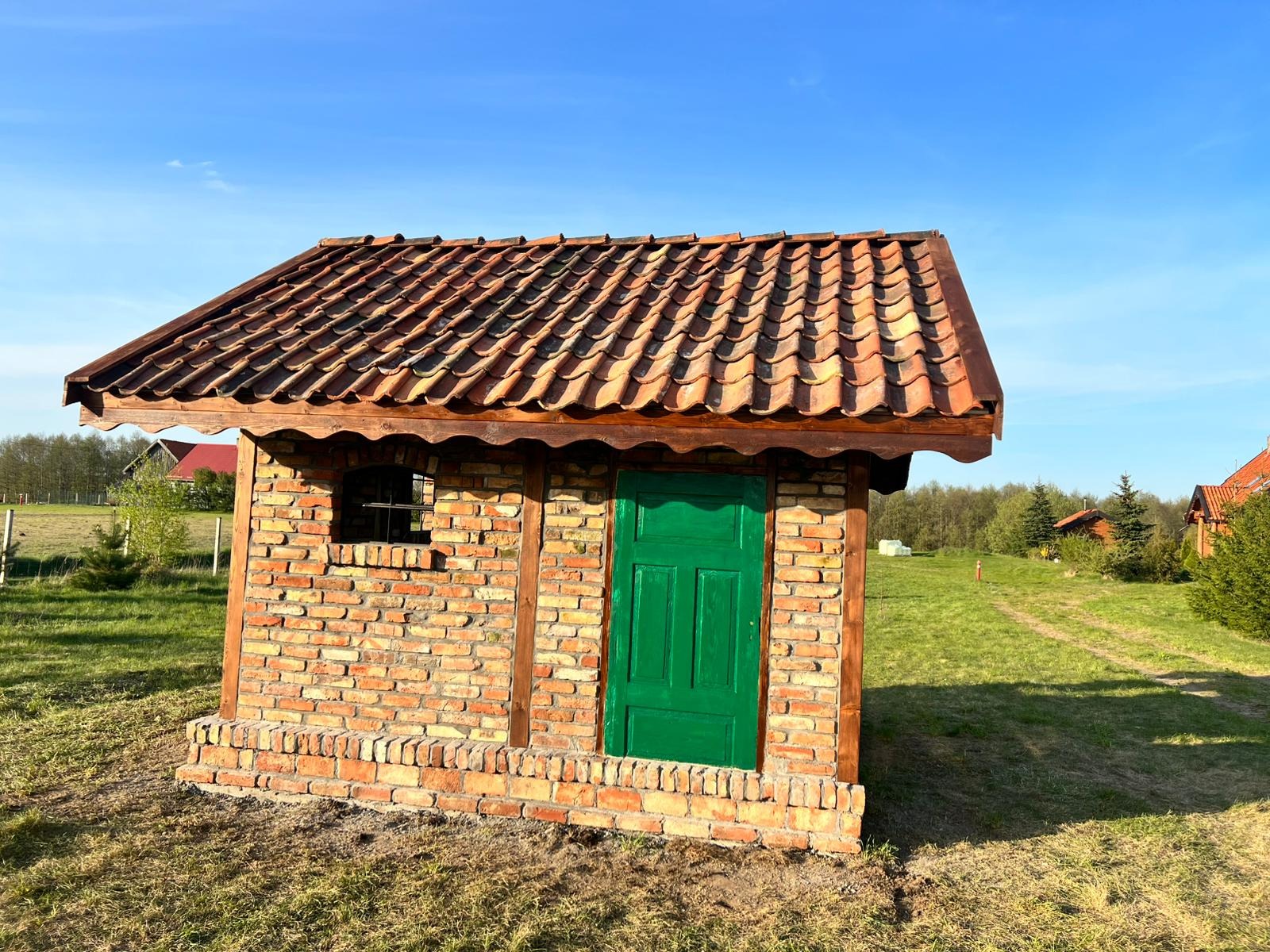 Mały budynek z cegły z zielonymi drzwiami i brązowym dachem z dachówki ceramicznej, otoczony trawnikiem i drzewami w oddali.