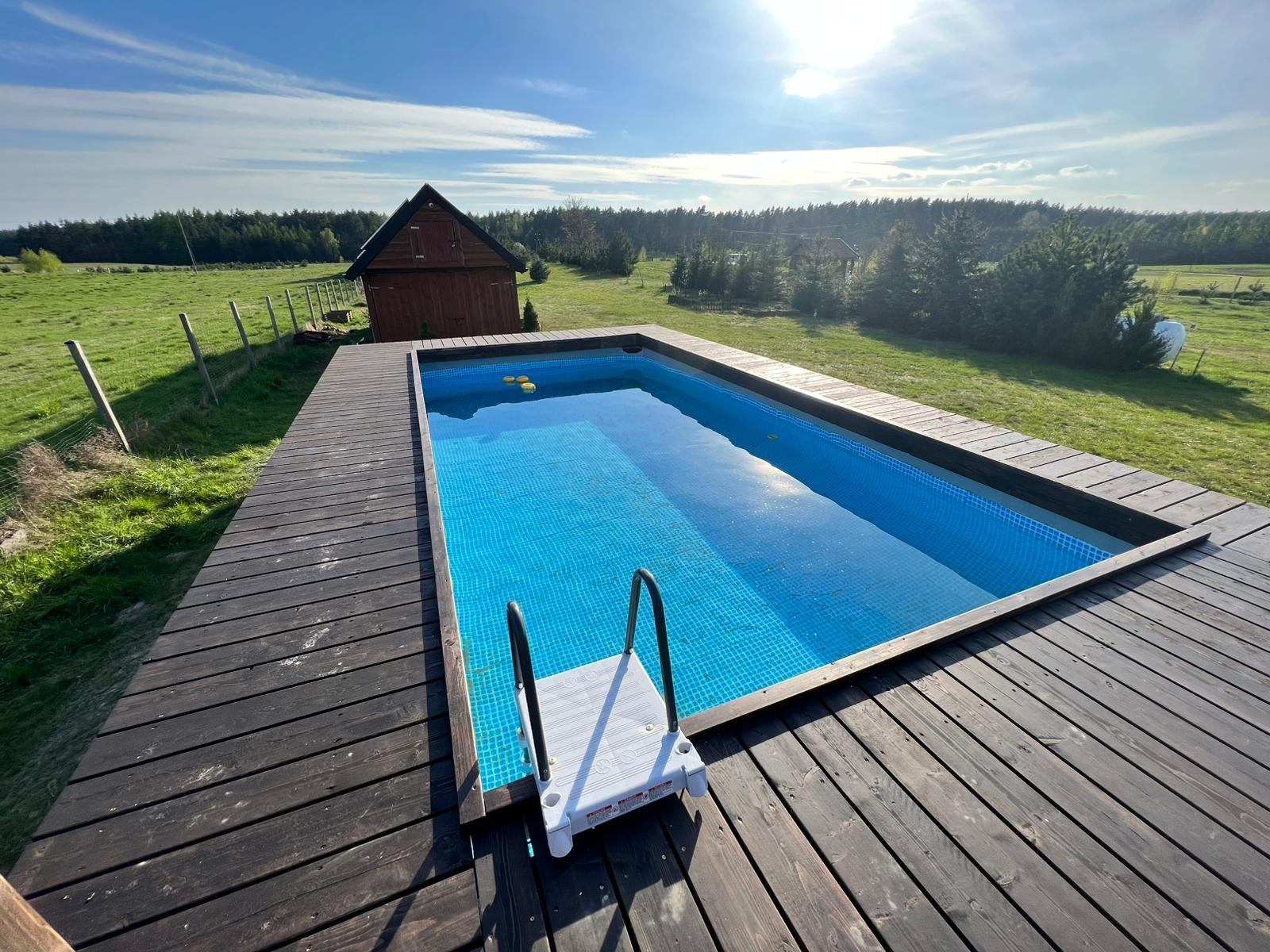 Prostokątny basen z niebieską mozaiką, otoczony drewnianym tarasem, z drabinką wejściową, na tle zielonego trawnika, lasu i błękitnego nieba.