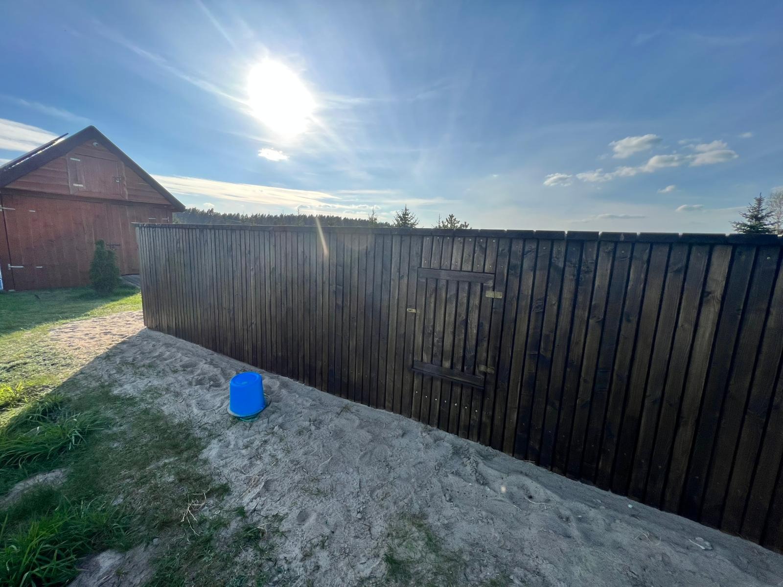 Ciemnobrązowe, drewniane ogrodzenie z furtką, widoczne od strony nasypu z piasku i trawy, w tle budynek gospodarczy i błękitne niebo z promieniami słońca.