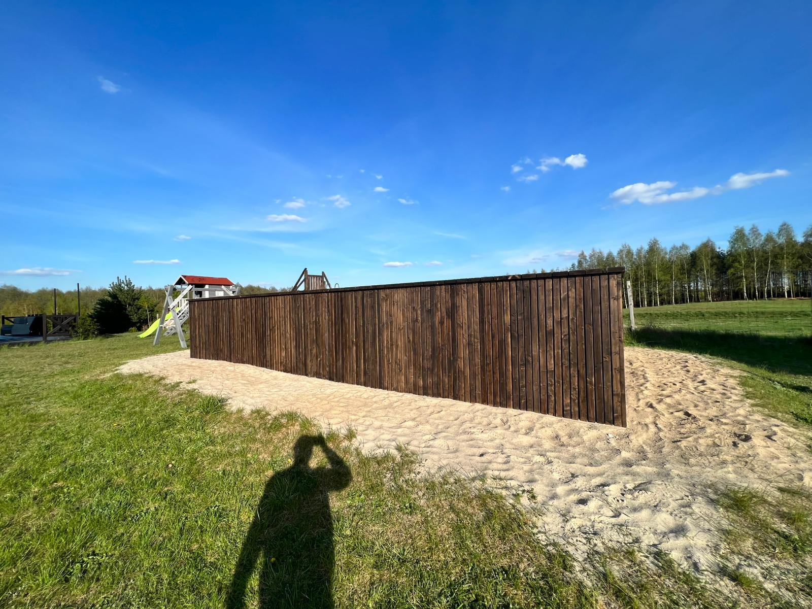 Długa, ciemna, drewniana ściana otaczająca obszar pokryty piaskiem, prawdopodobnie plac zabaw, z widocznym w tle domkiem i zjeżdżalnią oraz cieniem osoby robiącej zdjęcie.
