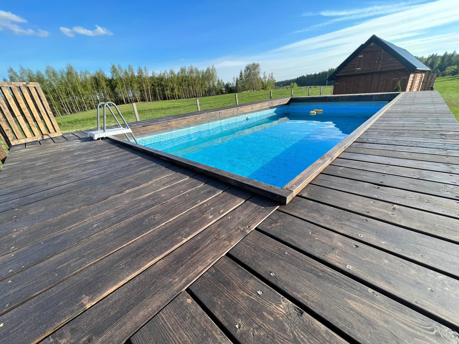 Niebieski basen z mozaiką, otoczony tarasem z ciemnego drewna, z drabinką i leżakiem, na tle zielonej łąki i lasu.