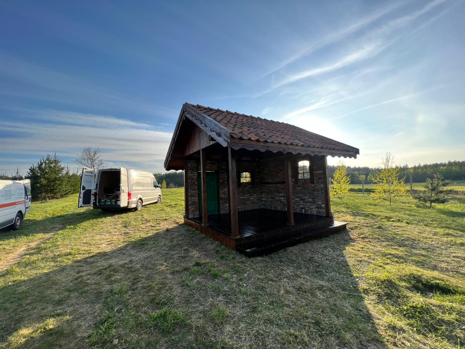 Gotowa altana z cegły i drewna z ciemną podłogą, z zielonymi drzwiami, na tle łąki i zaparkowanych samochodów dostawczych w słoneczny dzień.