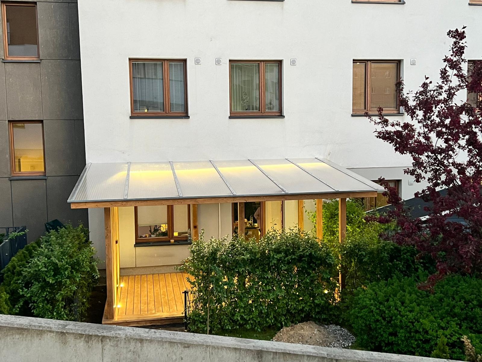 Zadaszony taras z drewnianą konstrukcją i przezroczystym dachem, oświetlony ciepłym światłem, przylegający do białego budynku mieszkalnego, otoczony zielenią.