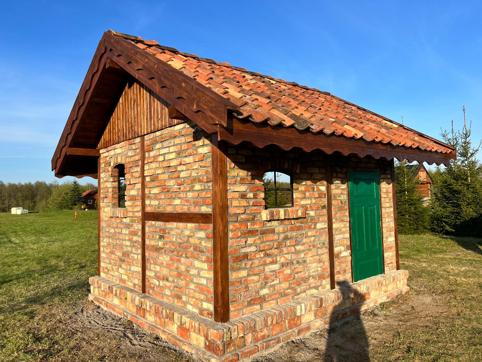 Mała, murowana konstrukcja z cegły z zielonymi drzwiami i oknami bez szyb, zwieńczona brązowym, drewnianym dachem z czerwonej dachówki, otoczona zielonym trawnikiem.