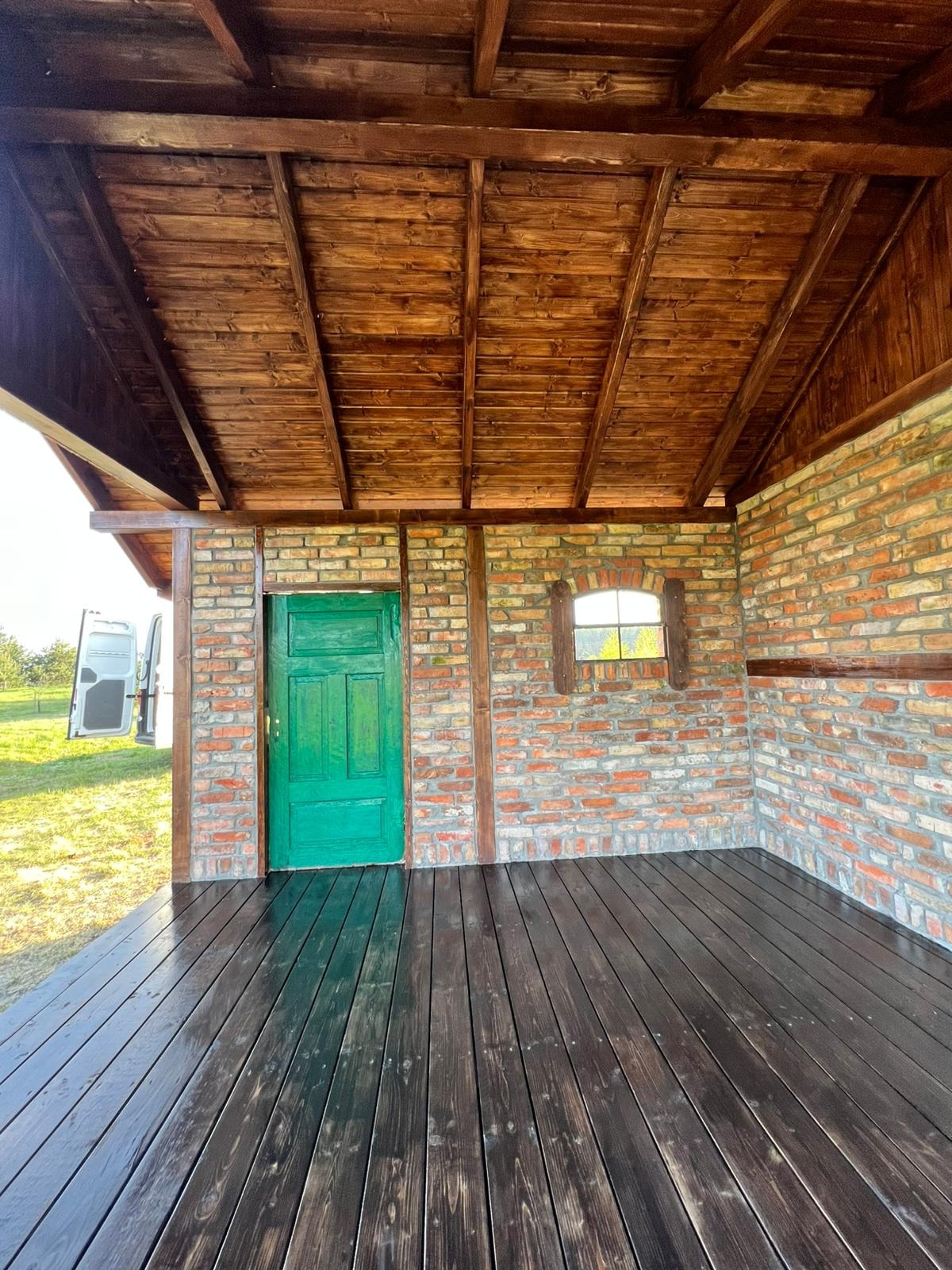 Zadaszona konstrukcja z ceglanymi ścianami, drewnianym sufitem i podłogą z desek, zielone drzwi, małe okno, widoczny samochód dostawczy w tle.