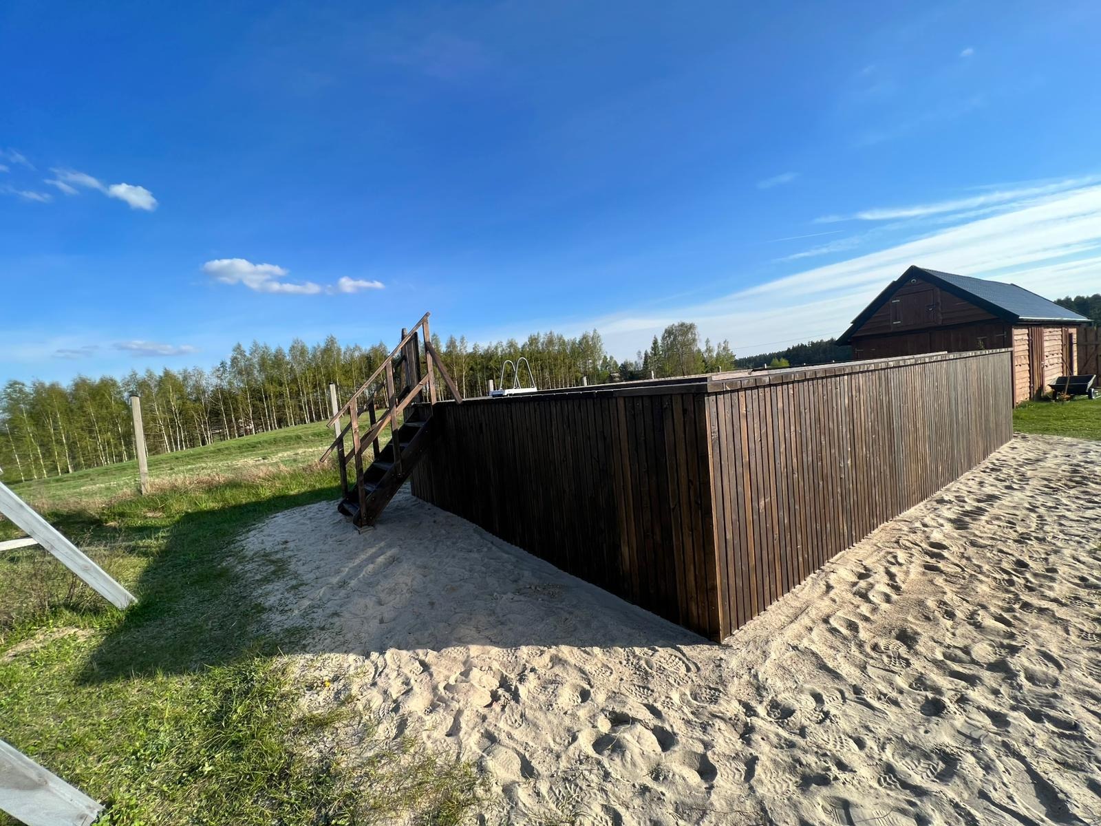 Drewniany taras z ciemnego drewna otoczony piaskiem, z widocznymi schodkami i budynkiem w tle, w słoneczny dzień.