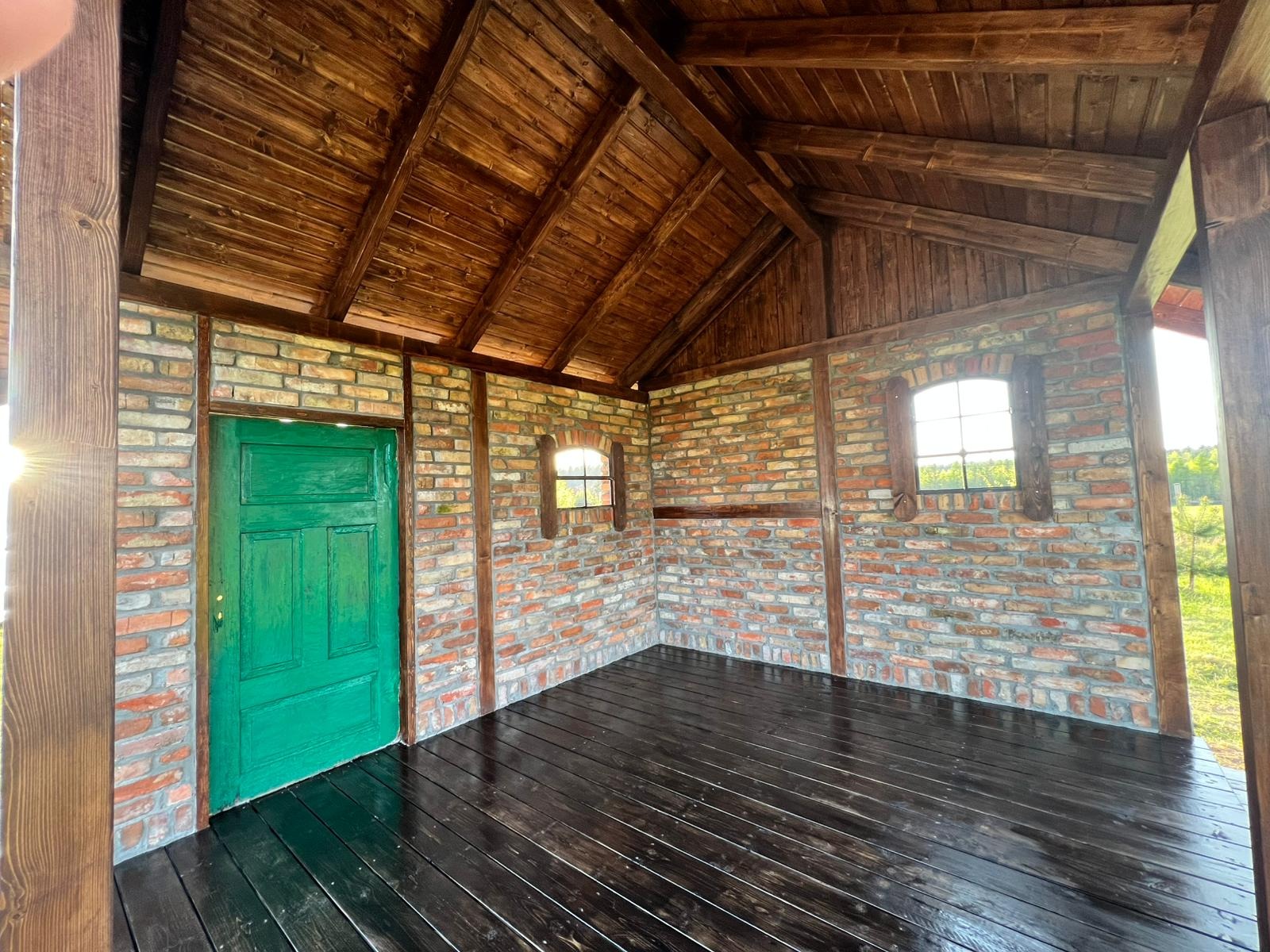 Wnętrze małej konstrukcji z ceglanymi ścianami, drewnianym dachem i podłogą, zielone drzwi po lewej stronie, dwa małe okna z drewnianymi ramami, widok z zewnątrz.