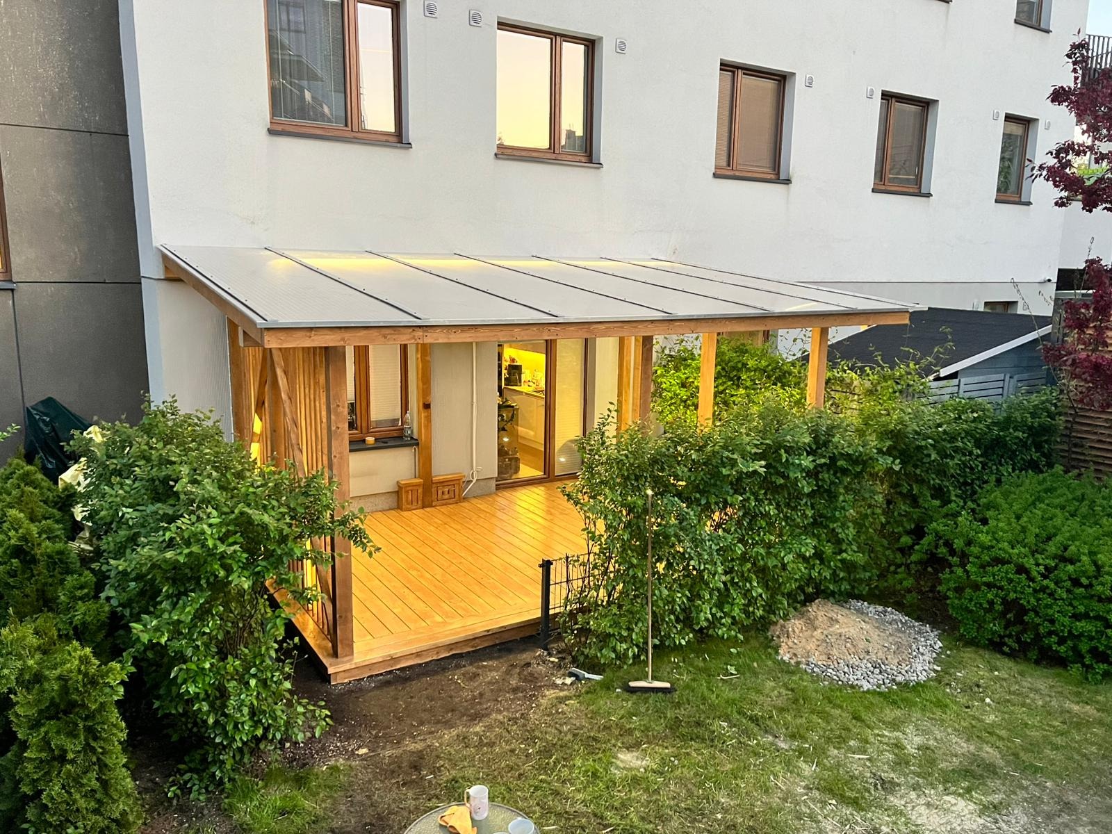 Zadaszony taras drewniany z przezroczystym dachem poliwęglanowym, przylegający do budynku mieszkalnego, otoczony zielenią, z widoczną stertą żwiru i narzędziami ogrodniczymi na trawniku.