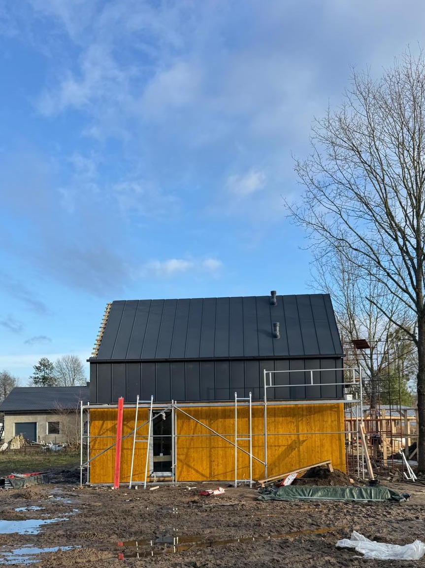 Wznoszony dom z drewnianą elewacją i ciemnym dachem na tle błękitnego nieba. Widoczne rusztowania i elementy konstrukcyjne. Etap budowy.