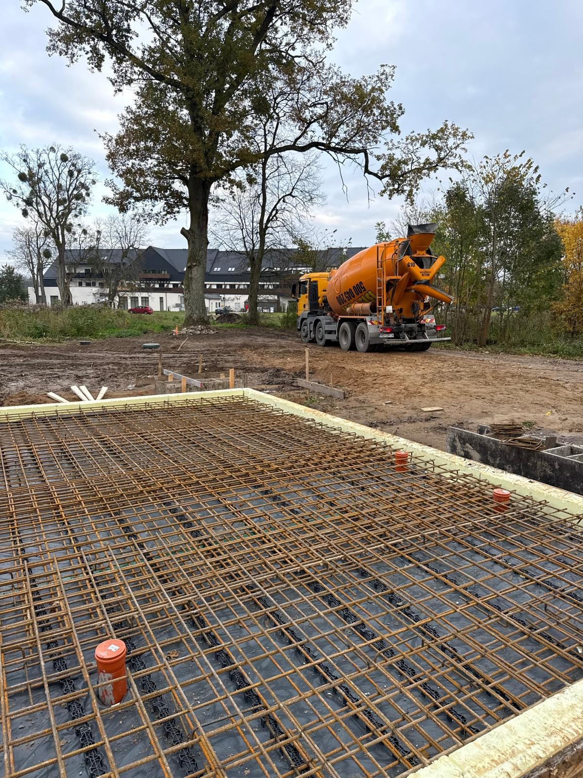 Zbrojenie fundamentu pod budowę, widoczna siatka z prętów stalowych na czarnej folii, obok stoi betoniarka. W tle budynek i drzewa.
