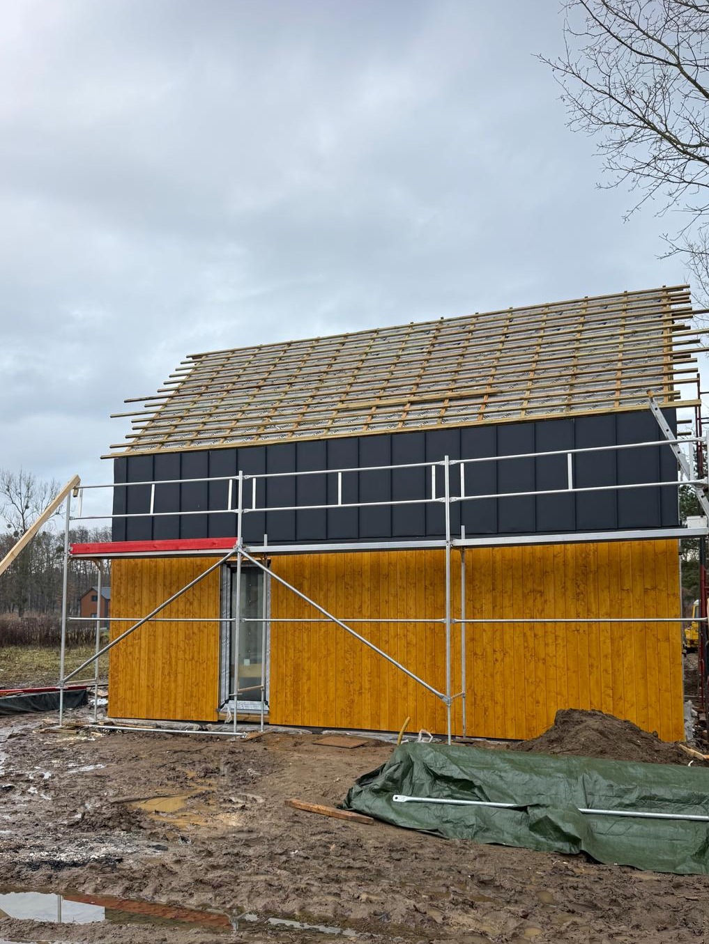 Budowa domu szkieletowego w Lubawie: żółta elewacja, czarne panele na dachu, rusztowania. Etap montażu łat dachowych na tle pochmurnego nieba.