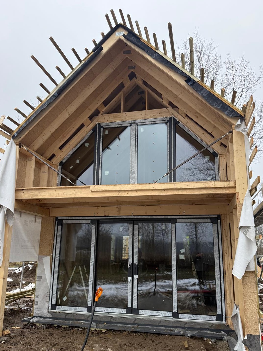 Surowy stan domu w konstrukcji szkieletowej z dużymi oknami, widoczne drewniane elementy konstrukcyjne i częściowo ośnieżone otoczenie. W tle drzewa bez liści.