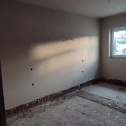 Pokój po tynkowaniu maszynowym, widoczne otwory na instalacje elektryczne, ślady po listwach przypodłogowych i zabrudzenia na podłodze.