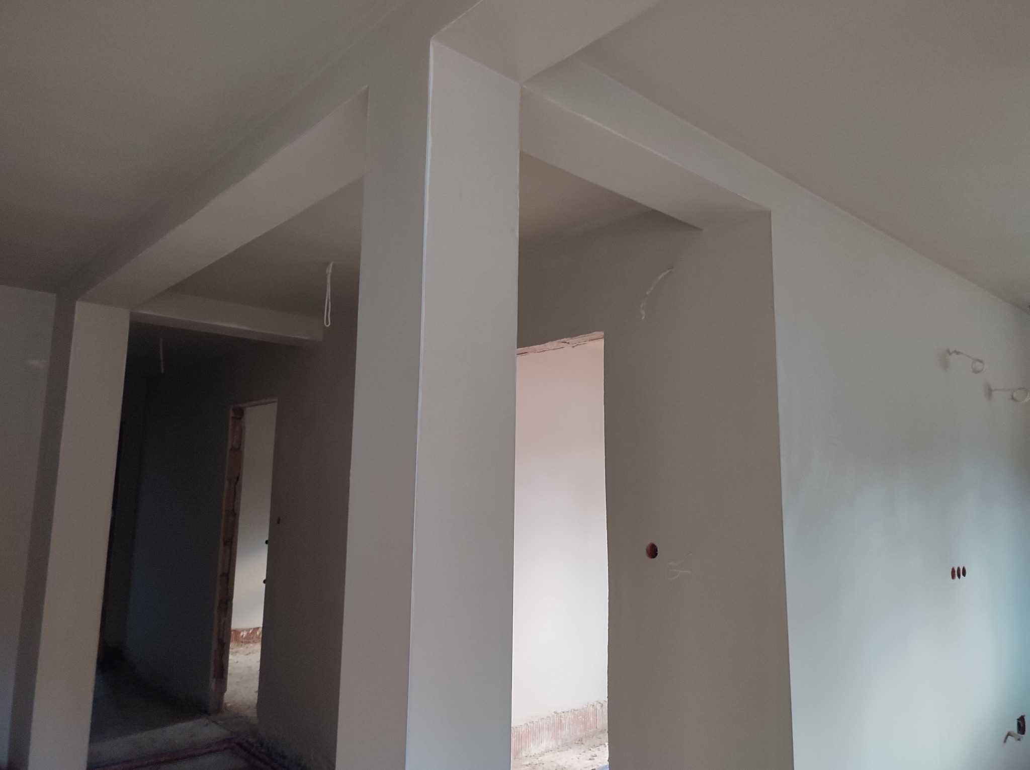 Wykończone, jasne wnętrze z pomalowanymi na biało ścianami i filarami, widoczne otwory drzwiowe prowadzące do kolejnych pomieszczeń w trakcie prac wykończeniowych.