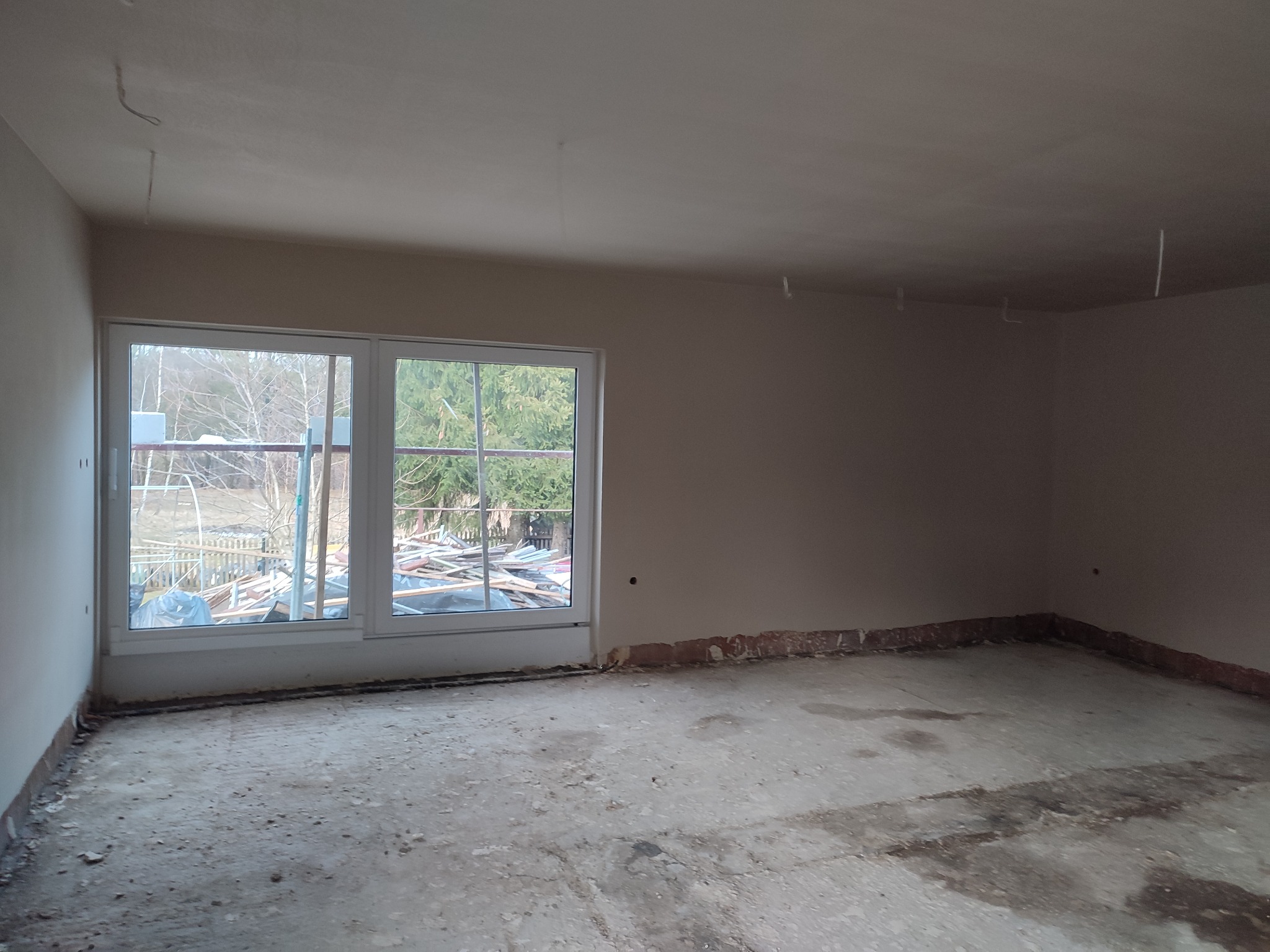 Pokój w trakcie remontu: ściany przygotowane pod tynkowanie, widoczne przewody elektryczne zwisające z sufitu, surowy beton na podłodze, okno z widokiem na podwórko z gruzem.