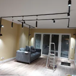 Zakończony montaż czarnego systemu oświetlenia szynowego w salonie z szarą sofą i drabiną, widoczne gniazdka elektryczne i radio.