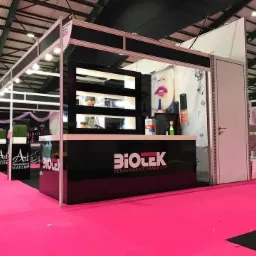 Stoisko targowe z czarnymi elementami i różową podłogą, prezentujące produkty do makijażu permanentnego, z oświetleniem sufitowym i widocznymi osobami w tle.