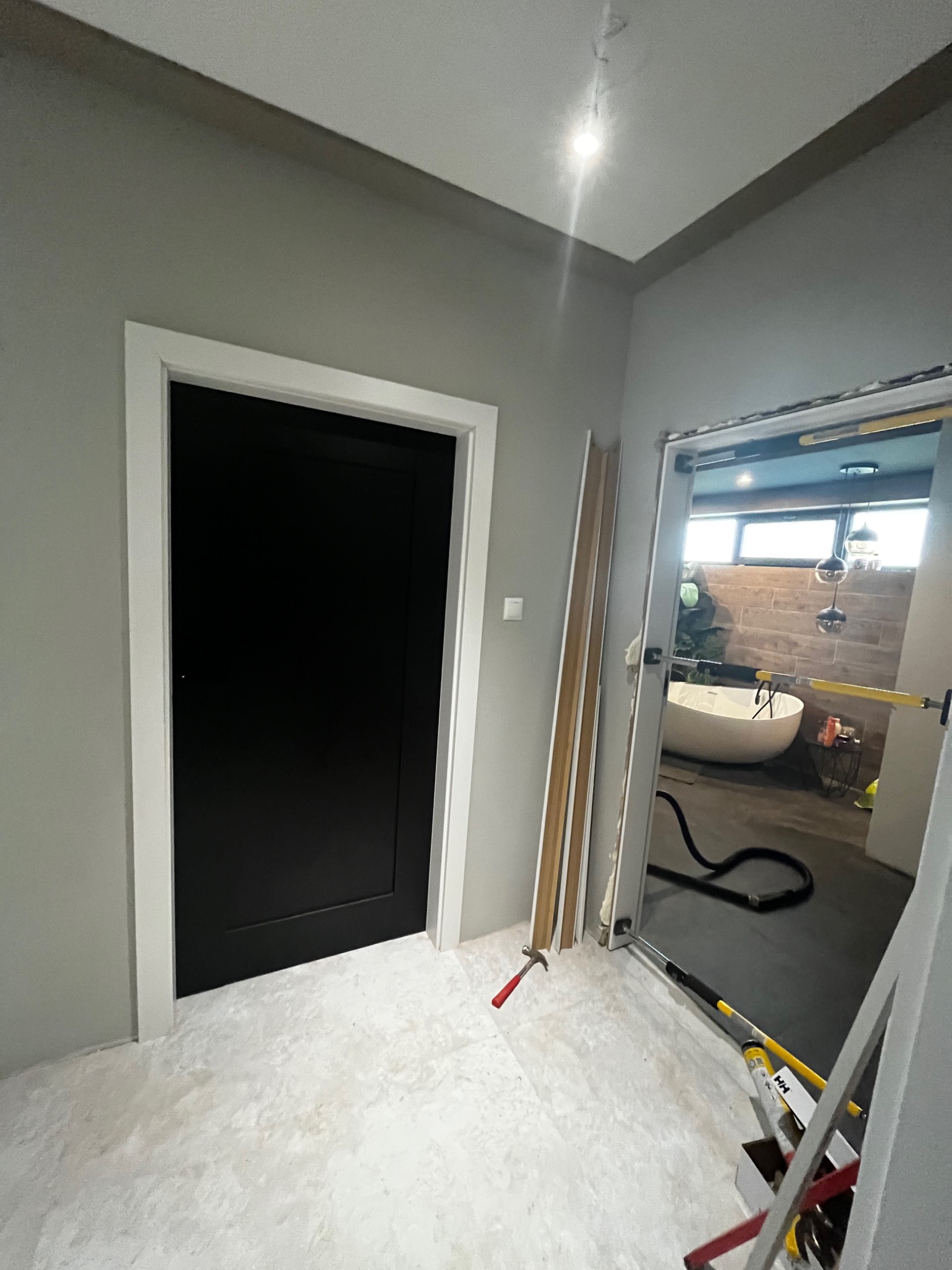 Ciemne drzwi z białym obramowaniem, widoczne narzędzia i przygotowania do montażu, w tle łazienka z wanną wolnostojącą.