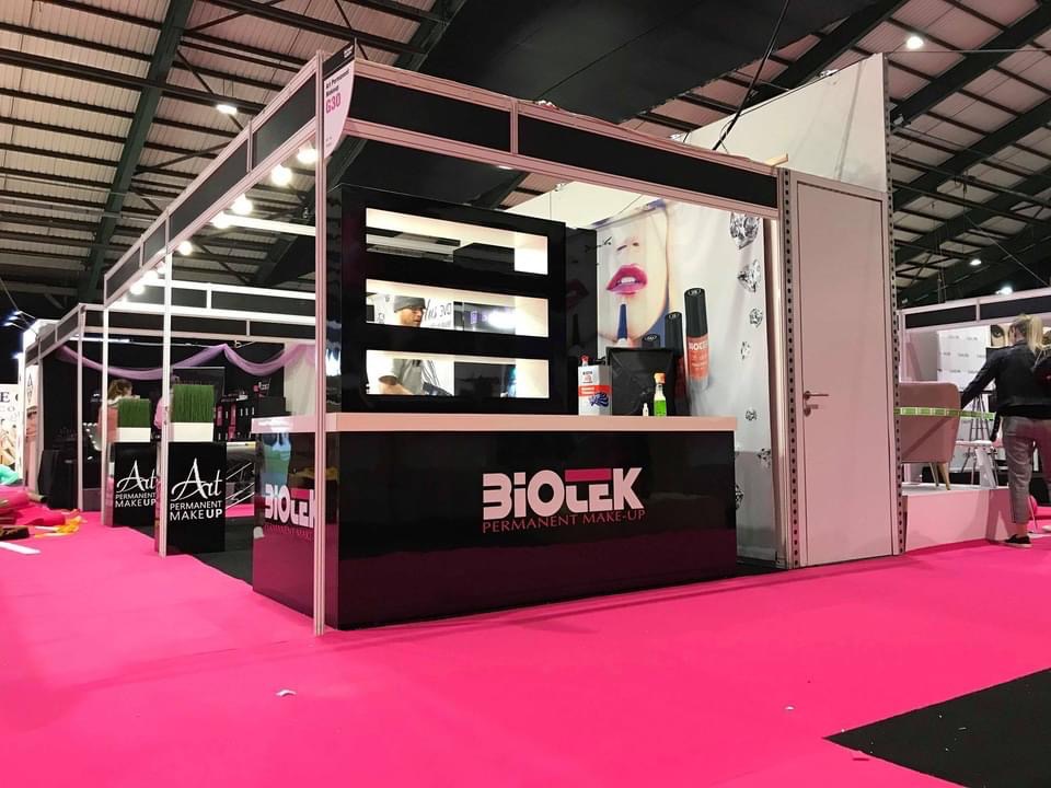 Stoisko targowe z czarnymi elementami i różową podłogą, prezentujące produkty do makijażu permanentnego, z oświetleniem sufitowym i widocznymi osobami w tle.