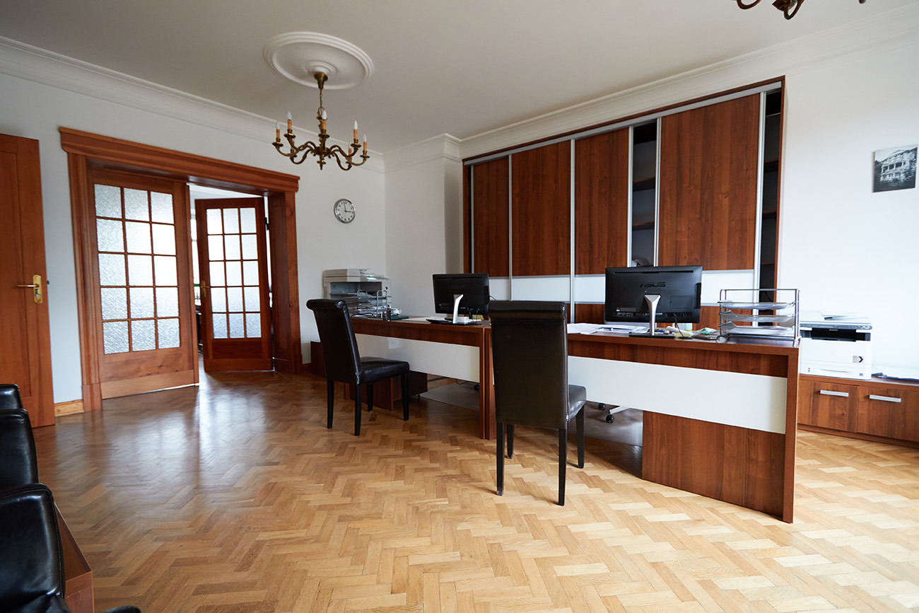 Jasne biuro z parkietem w jodełkę, biurkiem z dwoma stanowiskami komputerowymi, drewnianymi drzwiami z przeszkleniami i żyrandolem.