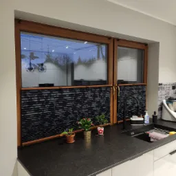 Ciemne, plisowane przesłony okienne w drewnianej ramie nad zlewem w kuchni, widoczne doniczkowe kwiaty i czarna bateria.