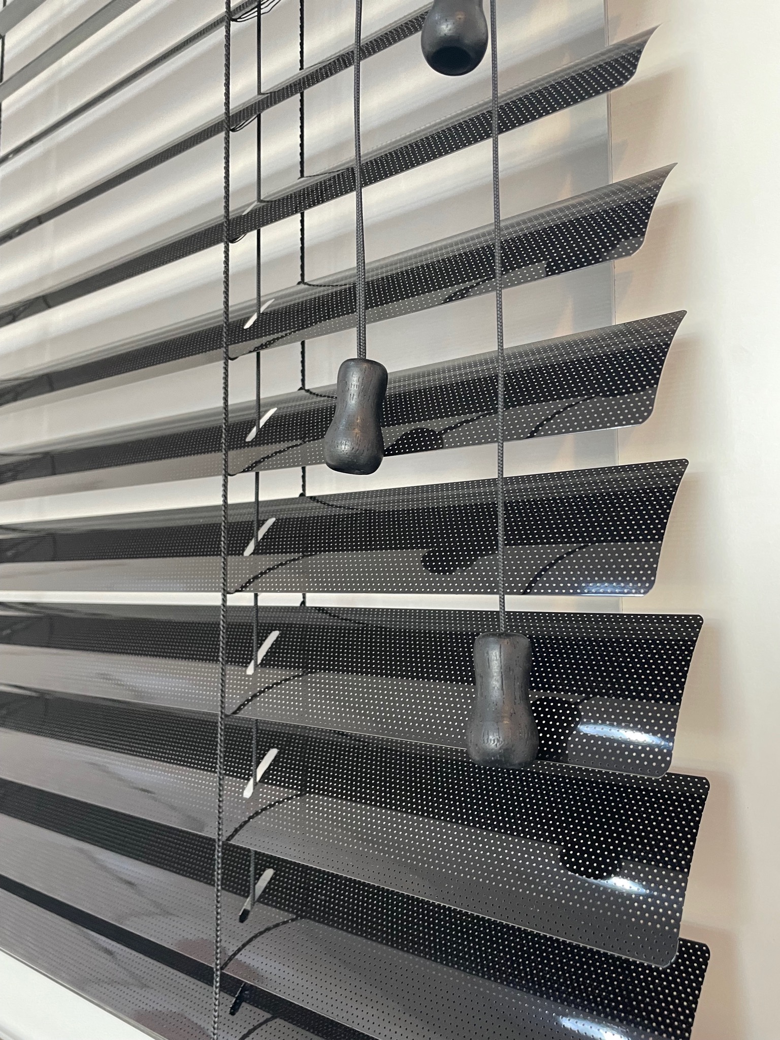 Częściowo zamknięte, perforowane, ciemne poziome osłony okienne z drewnianymi uchwytami sznurków.