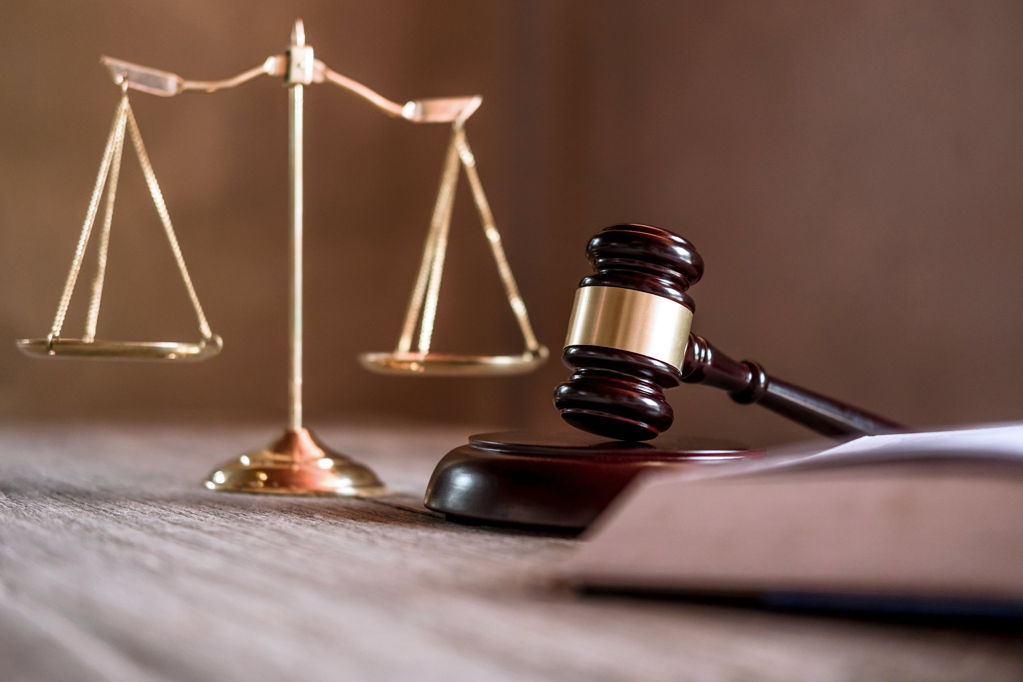 Złota waga sprawiedliwości w tle, z bliska drewniany młotek sędziowski na podstawie i fragment dokumentu na drewnianym stole.