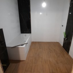 BUD-MASTER T&M SPÓŁKA Z OGRANICZONĄ ODPOWIEDZIALNOŚCIĄ - Remont łazienki w trakcie: ułożona podłoga z paneli imitujących drewno, białe płytki ścienne z czarnymi panelami dekoracyjnymi, sufit w trakcie szpachlowania, wanna obudowana płytkami.