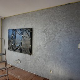 BUD-MASTER T&M SPÓŁKA Z OGRANICZONĄ ODPOWIEDZIALNOŚCIĄ - Ściana pokryta dekoracyjną, szarą strukturą, zabezpieczona folią i taśmą malarską w miejscu planowanego montażu telewizora. Na podłodze kartony ochronne, widoczna drabina aluminiowa.