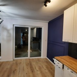 BUD-MASTER T&M SPÓŁKA Z OGRANICZONĄ ODPOWIEDZIALNOŚCIĄ - Nowoczesna kuchnia z białymi szafkami, drewnianym blatem, czarnym panelem ściennym i podłogą z paneli, widziana z perspektywy wnętrza mieszkania.