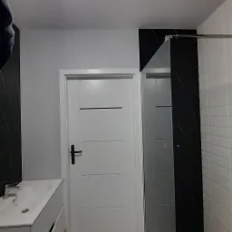 Nowoczesna łazienka po remoncie: biała umywalka z szafką, kabina prysznicowa z czarnym akcentem, białe drzwi z czarną klamką i wzorzysta podłoga.