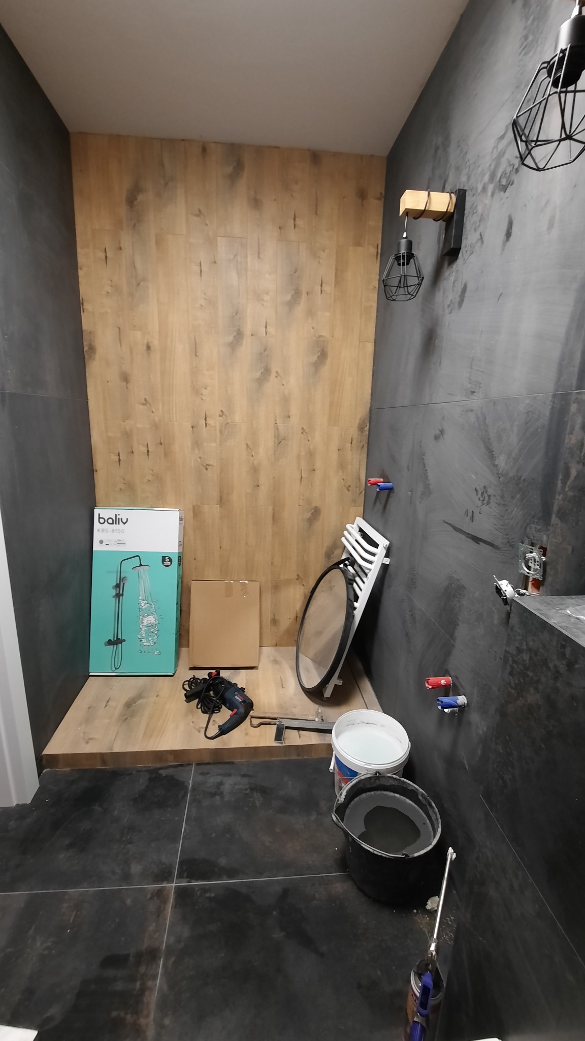 Wnętrze łazienki w trakcie wykańczania w Gdańsku: widoczne ciemne płytki na podłodze i ścianach, drewnopodobna okładzina na jednej ze ścian, przygotowane do montażu bateria prysznicowa w kartonie...