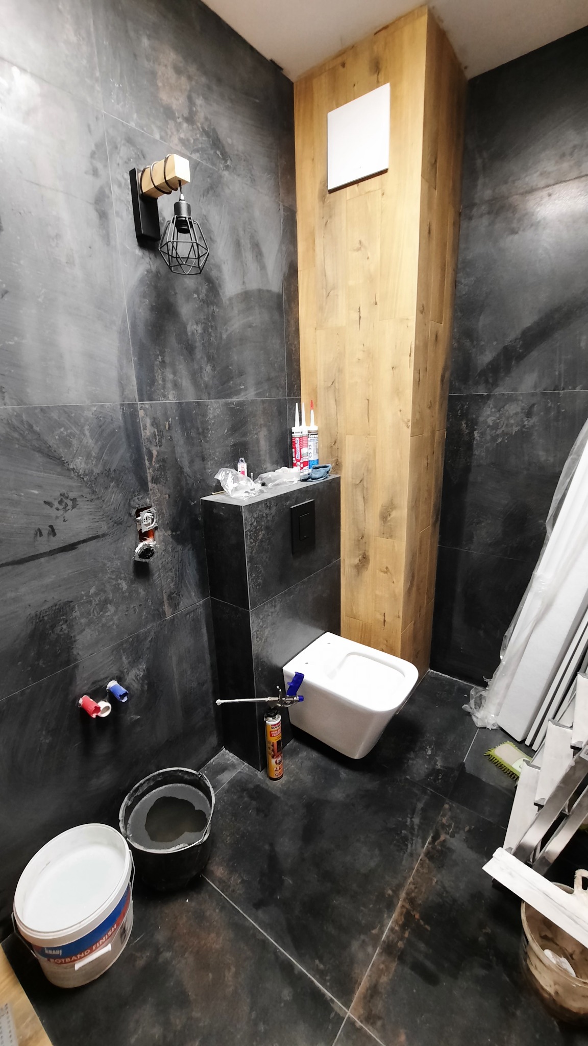 Wnętrze łazienki w trakcie prac wykończeniowych: widoczne czarne płytki imitujące kamień, podwieszana toaleta z zamontowaną armaturą, drewnopodobna obudowa pionu, wiadro z zaprawą, puszki silikonu...