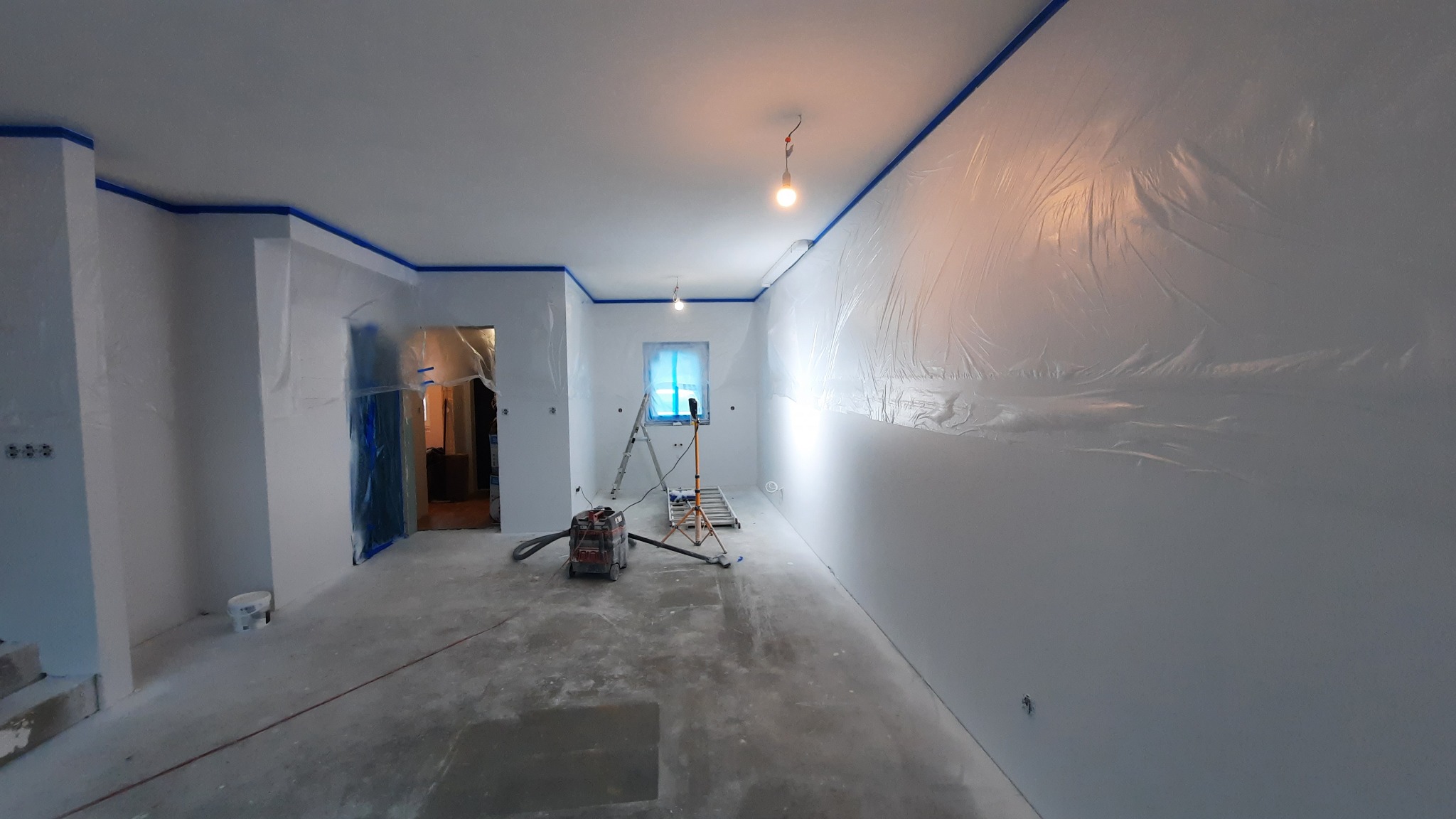 Pokój przygotowany do prac malarskich: ściany i sufit zabezpieczone folią, krawędzie oklejone taśmą malarską, widoczny odkurzacz przemysłowy, drabina i oświetlenie robocze.