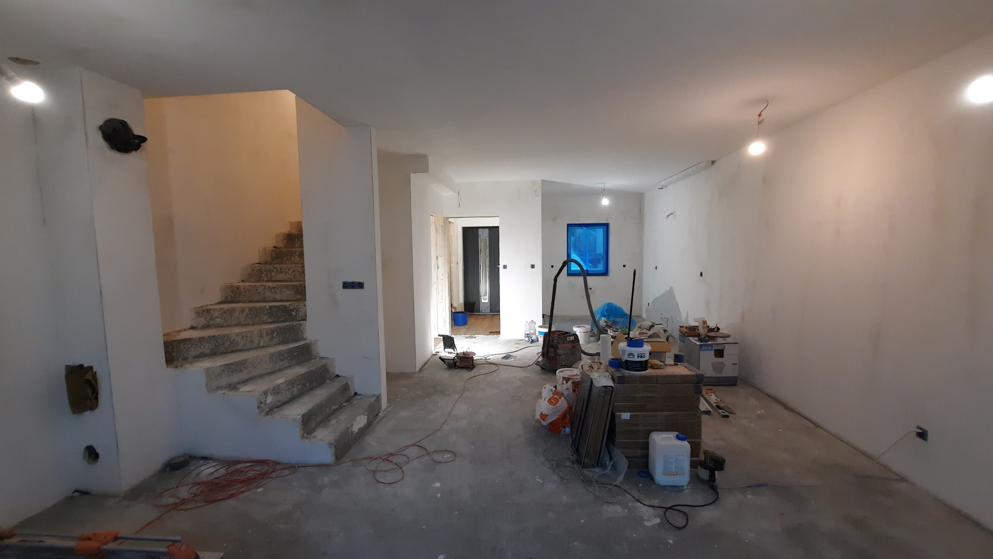 Wnętrze w trakcie prac wykończeniowych: surowe schody betonowe, świeżo otynkowane ściany, materiały budowlane i narzędzia rozstawione w pomieszczeniu.