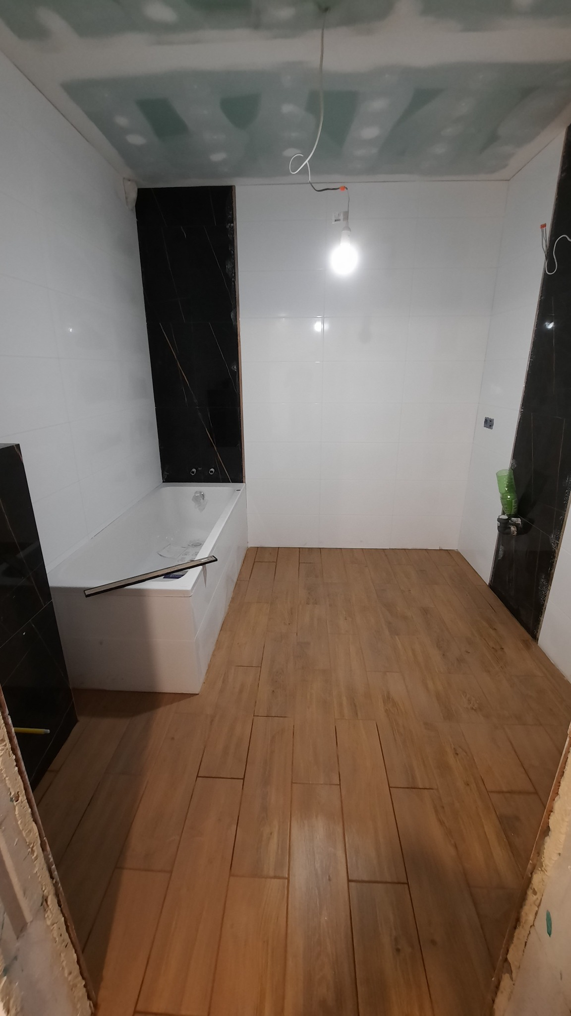 Remont łazienki w trakcie: ułożona podłoga z paneli imitujących drewno, białe płytki ścienne z czarnymi panelami dekoracyjnymi, sufit w trakcie szpachlowania, wanna obudowana płytkami.