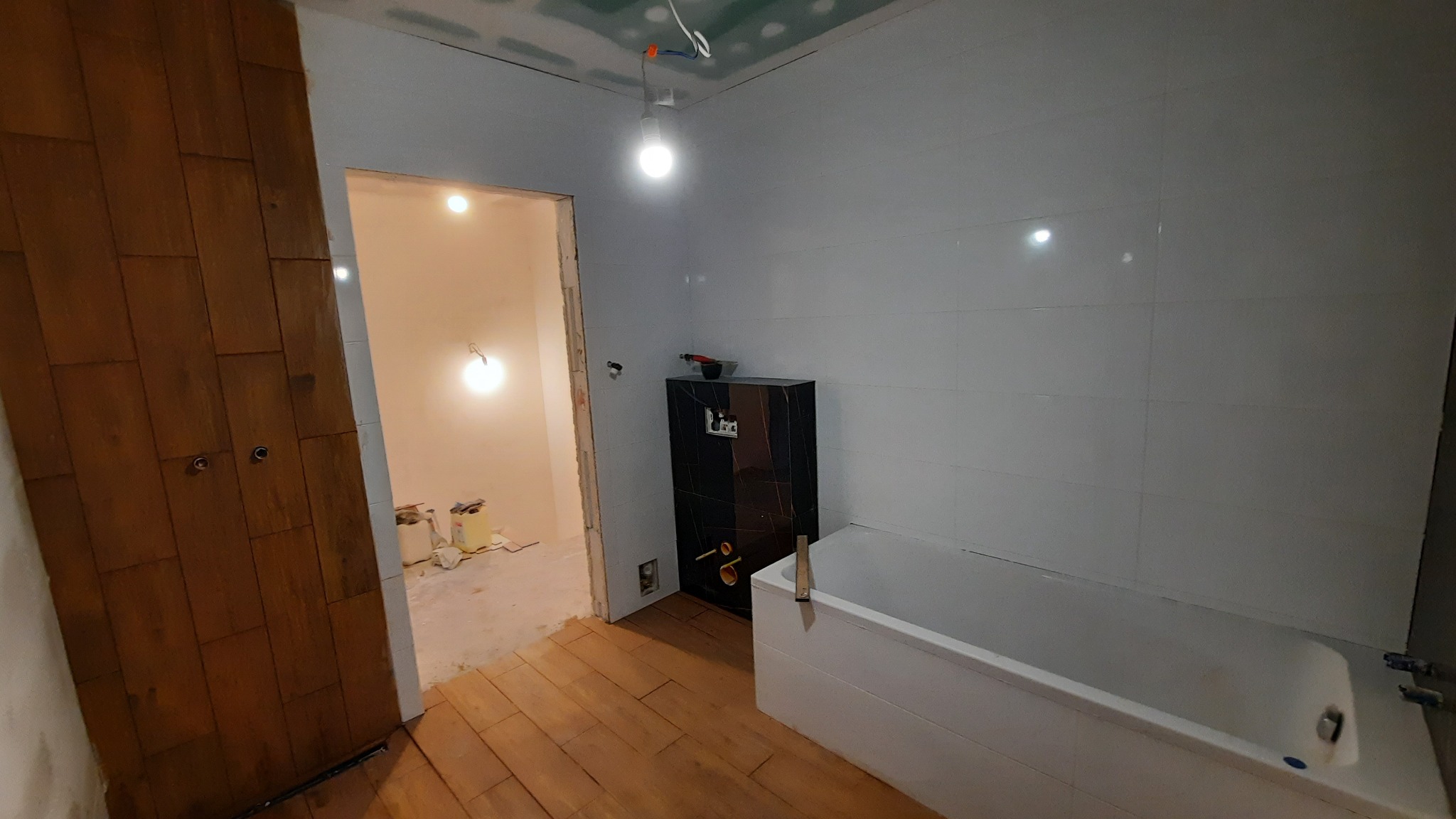 Łazienka w trakcie remontu: białe płytki na ścianach, zabudowa podtynkowego WC w czarnym marmurze, biała wanna, sufit z płyt karton-gips, drewniana podłoga i drzwi.