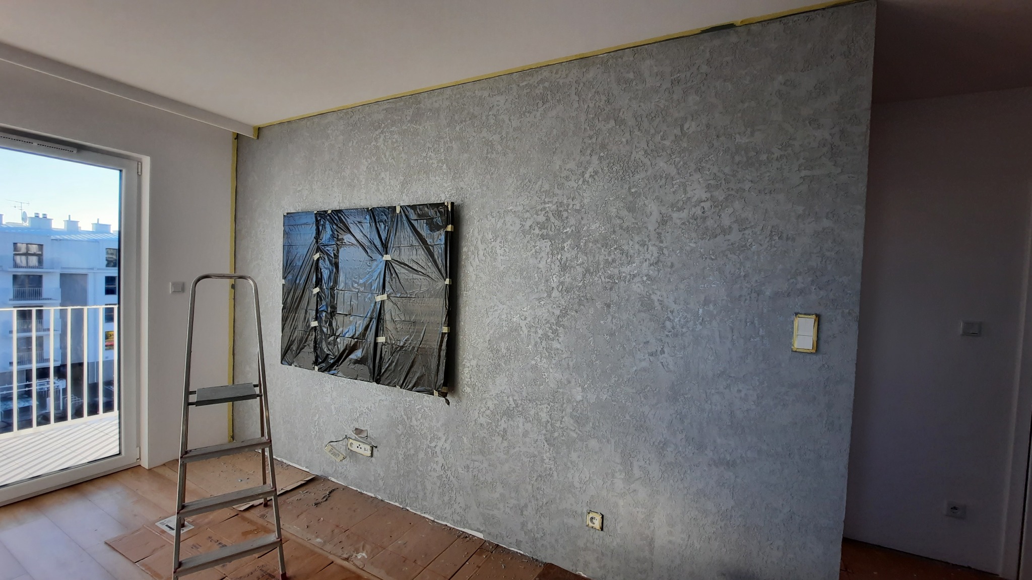 Ściana pokryta dekoracyjną, szarą strukturą, zabezpieczona folią i taśmą malarską w miejscu planowanego montażu telewizora. Na podłodze kartony ochronne, widoczna drabina aluminiowa.
