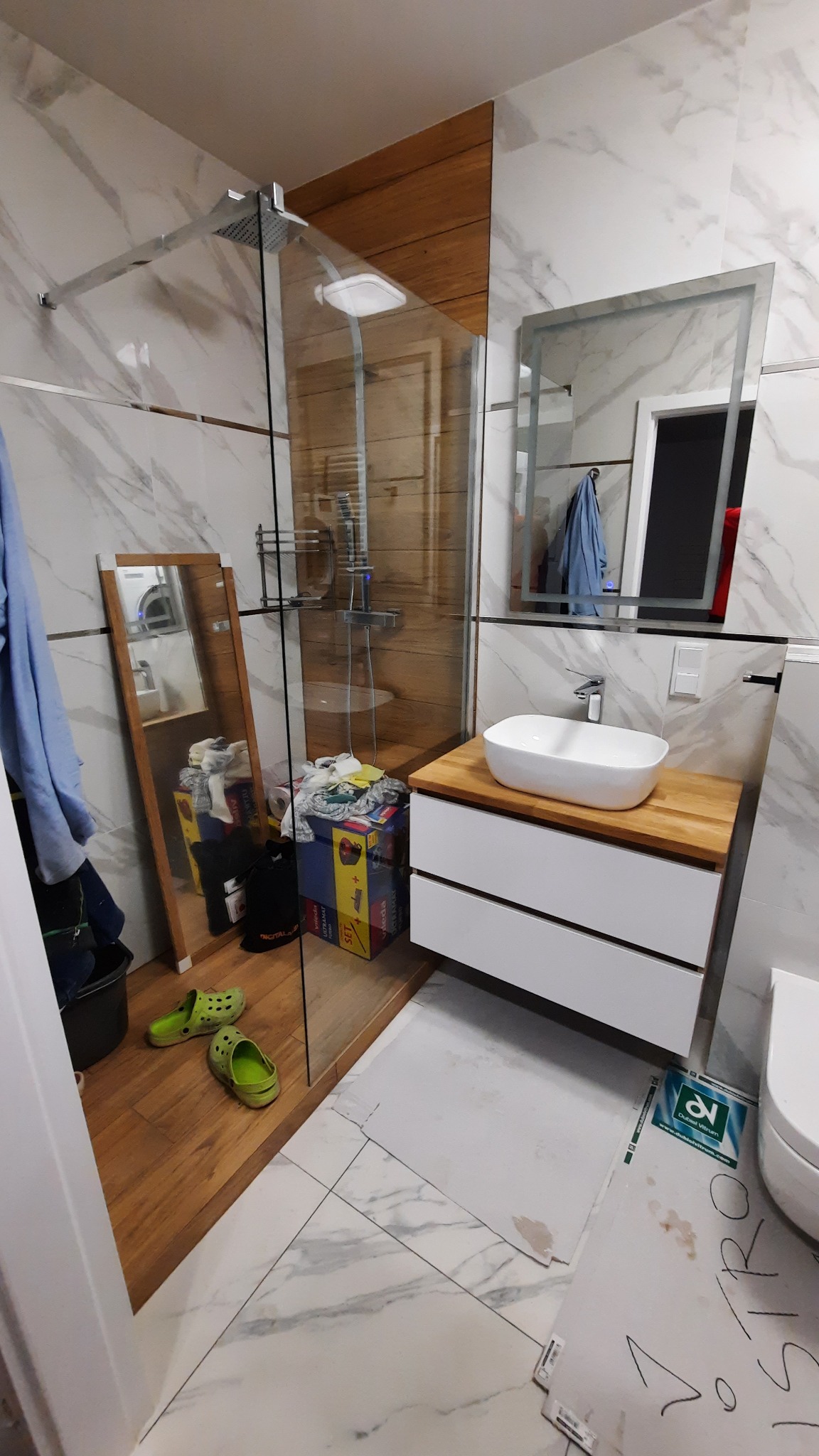 Nowoczesna łazienka z kabiną prysznicową typu walk-in, umywalką nablatową na drewnianym blacie i marmurowymi płytkami na ścianach, w trakcie wykańczania, widoczne kartony i niedbale pozostawione...