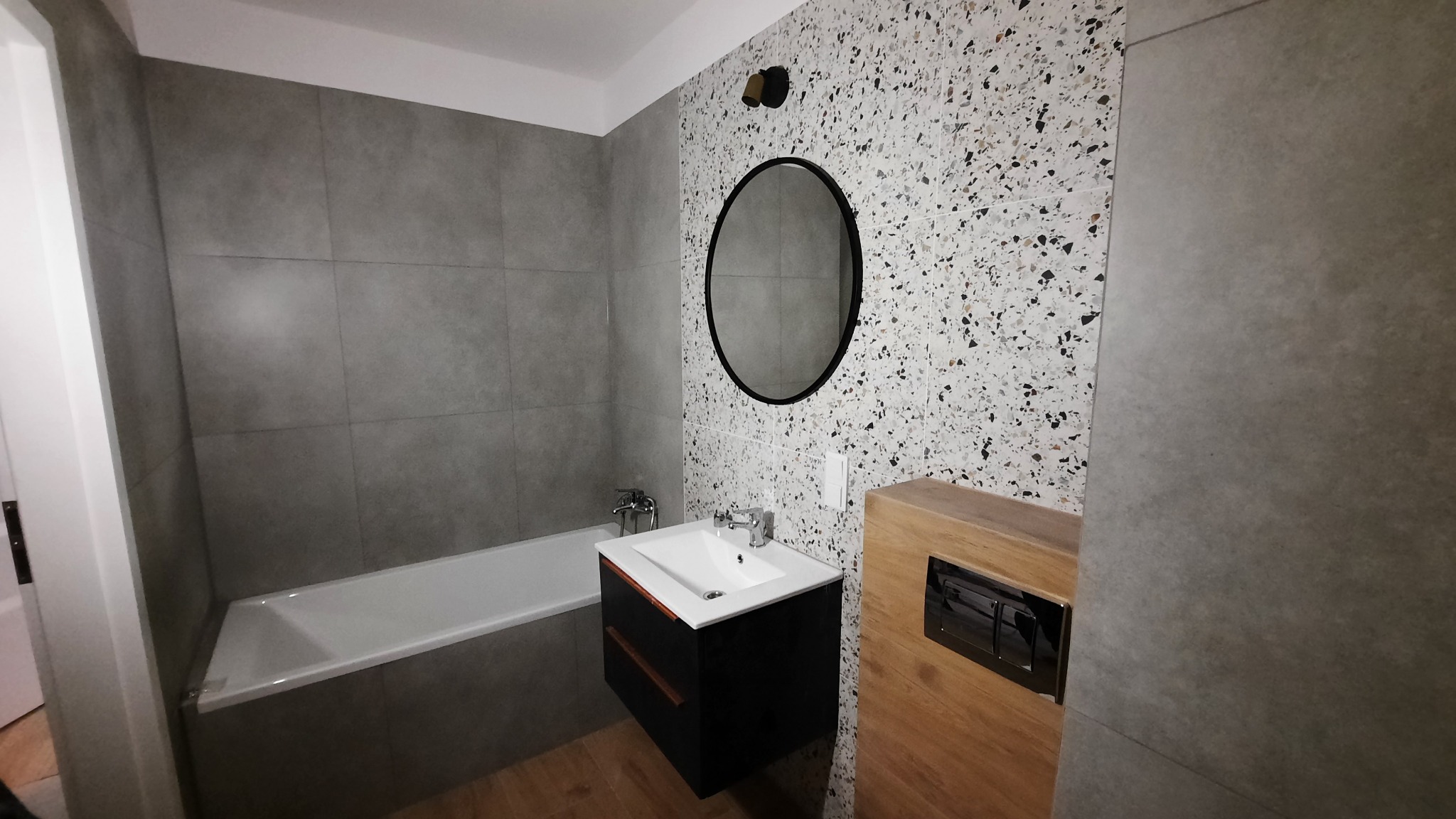 Nowoczesna łazienka z szarymi i terazzo płytkami, czarną szafką pod umywalką z drewnianymi akcentami oraz okrągłym lustrem.