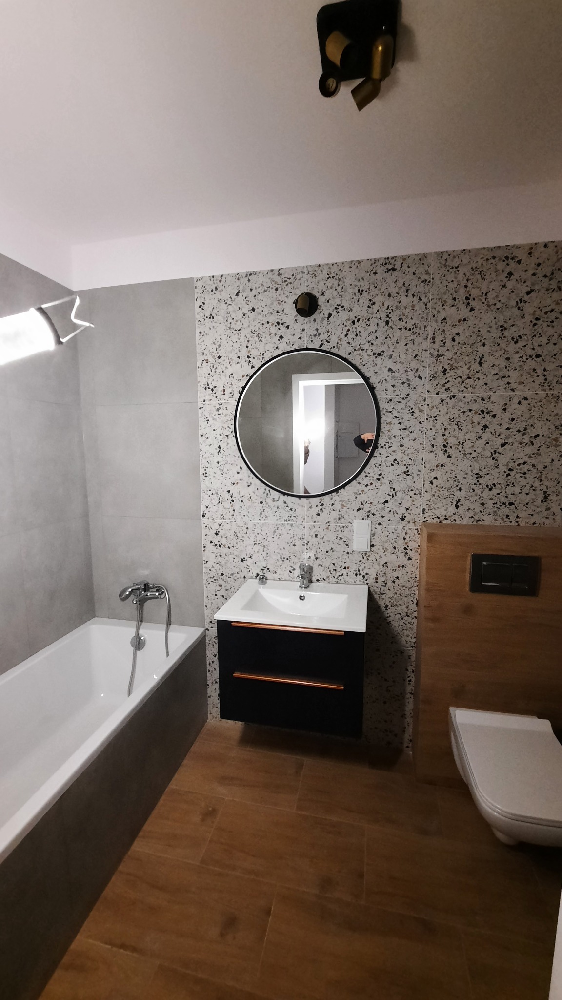 Nowoczesna łazienka z płytkami imitującymi lastryko na ścianie, okrągłym lustrem, czarną szafką pod umywalką z miedzianymi uchwytami, drewnopodobnymi płytkami na podłodze i minimalistyczną armaturą.