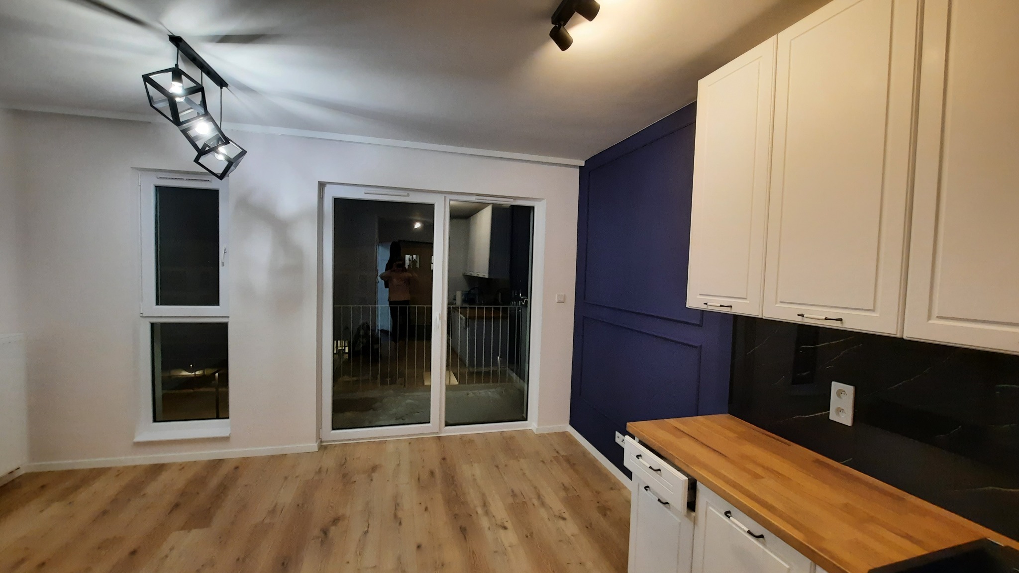 Nowoczesna kuchnia z białymi szafkami, drewnianym blatem, czarnym panelem ściennym i podłogą z paneli, widziana z perspektywy wnętrza mieszkania.