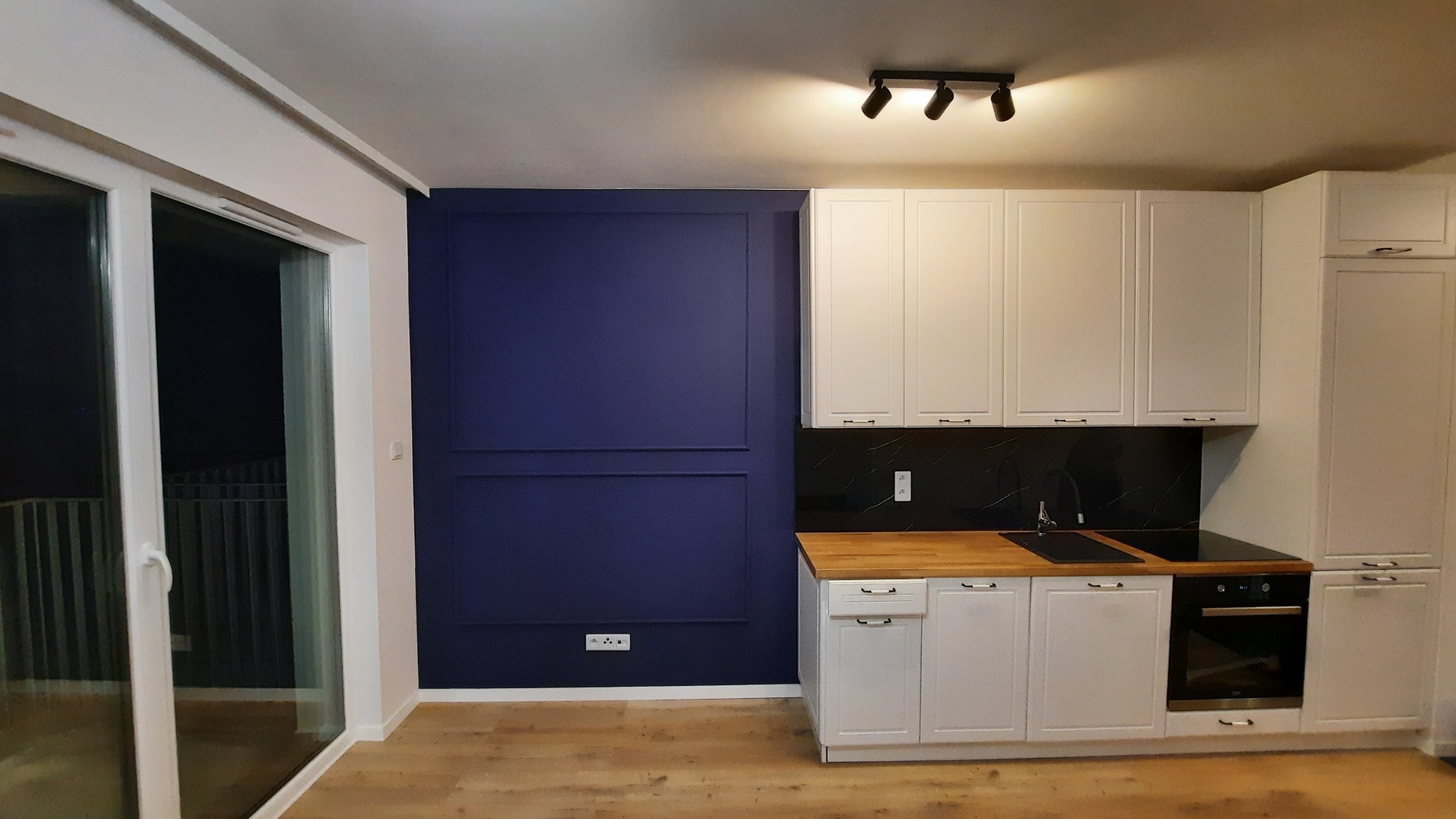 Nowoczesna kuchnia z białymi szafkami, drewnianym blatem i granatową ścianą z dekoracyjnymi listwami, oświetlona trzema reflektorami punktowymi.