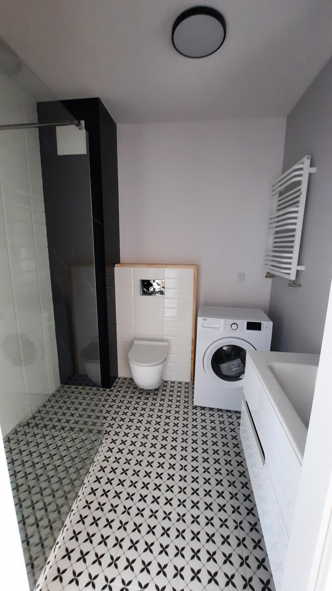 Nowoczesna łazienka w odcieniach bieli i szarości z wzorzystą podłogą, widoczny fragment kabiny prysznicowej, podwieszana toaleta, pralka i grzejnik drabinkowy.