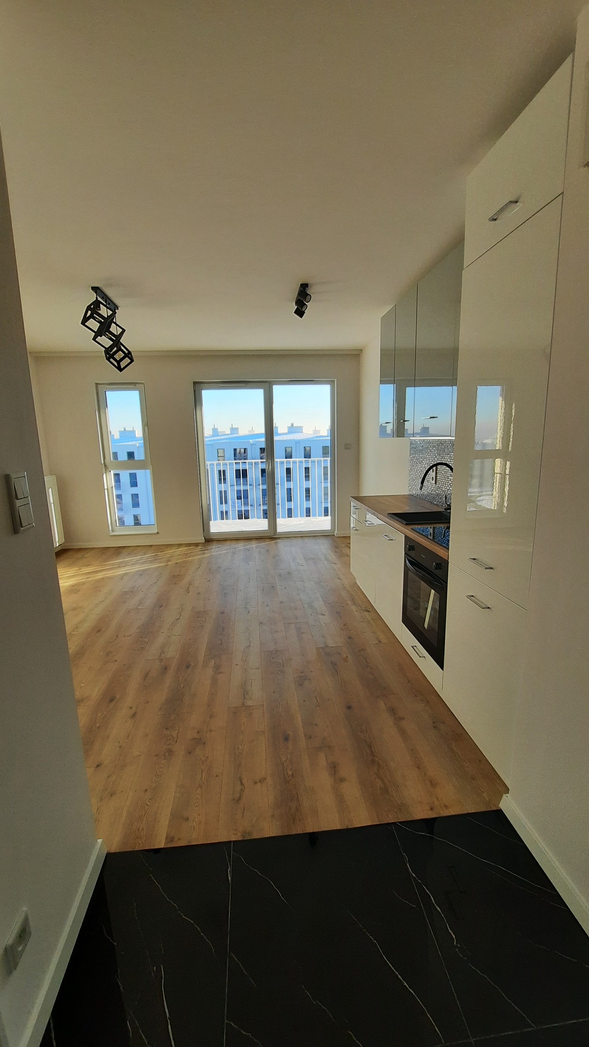 Jasne, przestronne wnętrze mieszkania po remoncie: drewniane panele podłogowe, biała zabudowa kuchenna z czarnym blatem i oknem balkonowym z widokiem na miasto.
