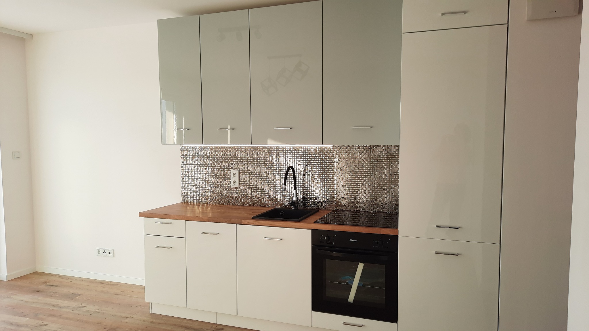 Nowoczesna kuchnia z białymi szafkami, drewnianym blatem i mozaiką nad blatem, czarny zlew i płyta indukcyjna.