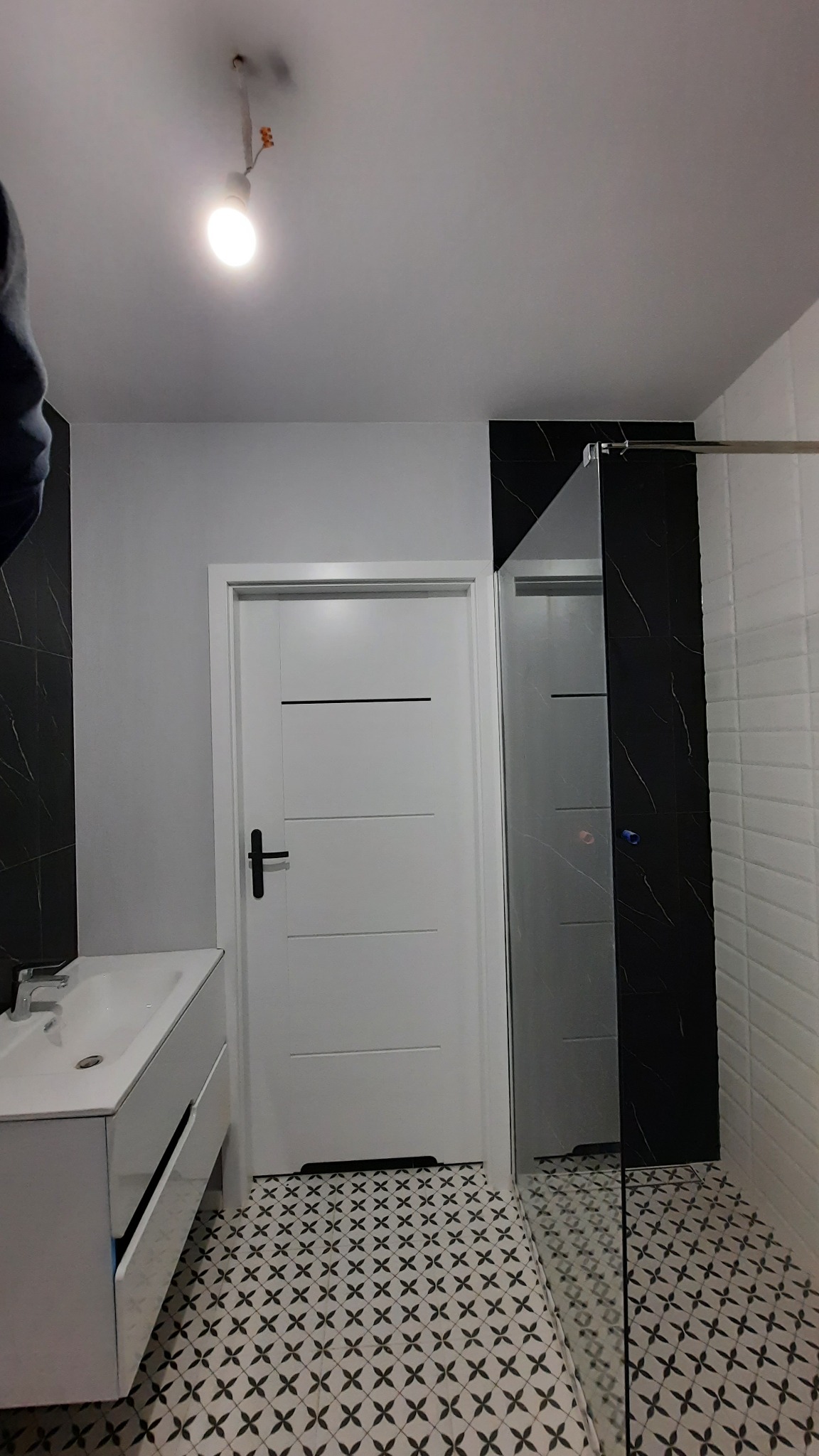 Nowoczesna łazienka po remoncie: biała umywalka z szafką, kabina prysznicowa z czarnym akcentem, białe drzwi z czarną klamką i wzorzysta podłoga.