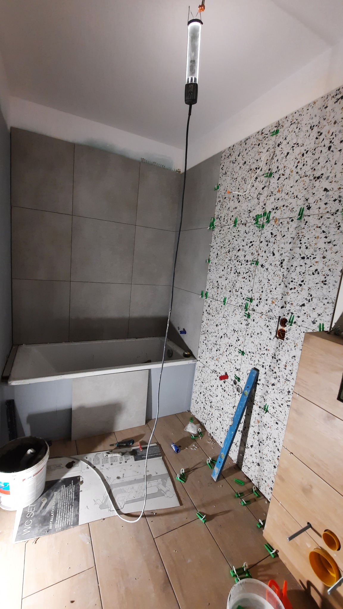 Łazienka w trakcie wykańczania: szare płytki na ścianie nad wanną, mozaika na drugiej ścianie z plastikowymi krzyżykami dystansowymi, poziomica oparta o ścianę, wiadro z zaprawą i narzędzia...