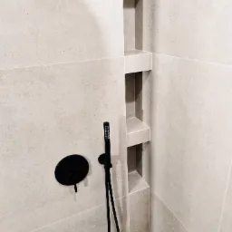 Minimalistyczna kabina prysznicowa z beżowymi płytkami i czarną armaturą, wnęki w ścianie. Nowoczesny design, stonowana kolorystyka, precyzyjne wykończenie.